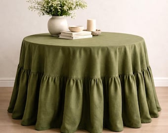 Op de afbeelding: Een olijfgroene ronde tafelkleed met een gefranjerde rand. De tafel is gedekt met een witte vaas met bloemen, boeken, een kleine kom en een kaars. Het tafelkleed is gemaakt van een textuurstof.