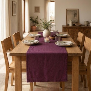 May include: A rectangular wooden dining table featuring a deep purple linen table runner. The table is set with four white plates, each topped with a folded napkin and a small green sprig. A white vase with greenery and a candle sit in the centre.