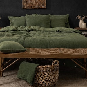 Può includere: Una camera da letto con biancheria da letto verde oliva, tra cui un copripiumino e cuscini. Una panca in legno è ai piedi del letto e sotto c'è un cesto di vimini. A sinistra ci sono una lampada e un comodino.