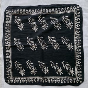 May include: A black square bandana with a white floral and leaf pattern. The bandana has a decorative border with a repeating geometric design. The floral pattern is evenly spaced across the center of the bandana.