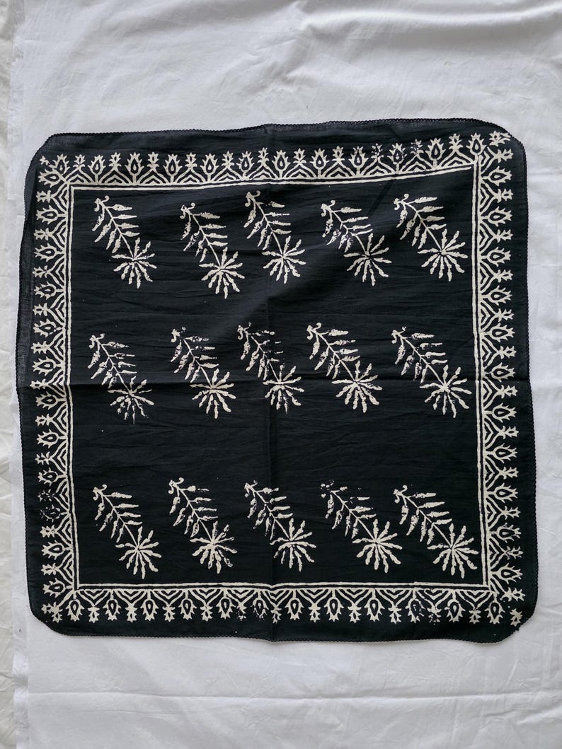 May include: A black square bandana with a white floral and leaf pattern. The bandana has a decorative border with a repeating geometric design. The floral pattern is evenly spaced across the center of the bandana.