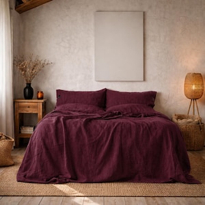 May include: A bedroom with a bed dressed in deep burgundy linen bedding. The room has a neutral colour palette with a wooden bedside table, a woven lamp, and a large blank canvas on the wall. A woven basket and dried flowers add to the cosy atmosphere.