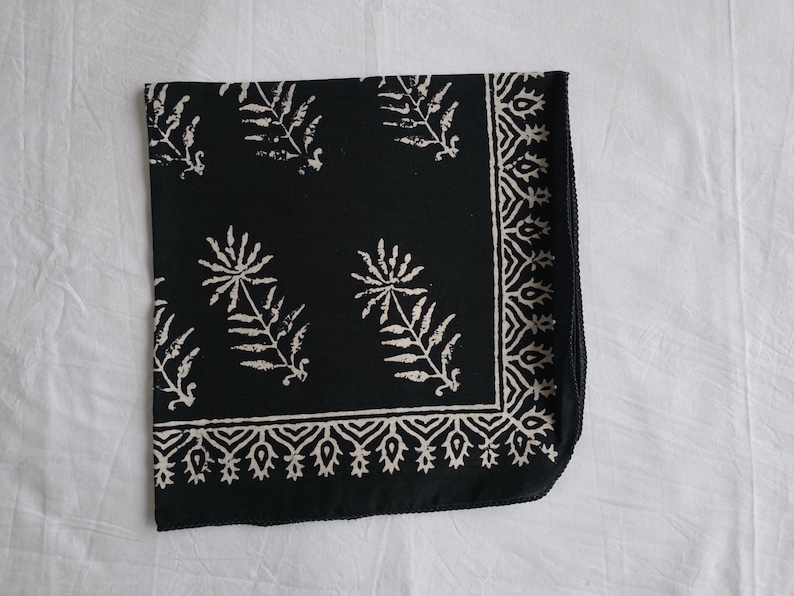May include: A folded black square textile with a white floral and leaf pattern. The design includes stylized flowers and leaves, with a decorative border along one edge. The textile is likely cotton or linen.