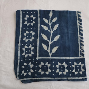 May include: A square, indigo-dyed textile with a white floral and leaf pattern. The design includes a central leaf motif and a border with star-like shapes. The fabric is folded and lies on a white surface.