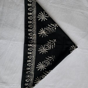 May include: Black bandana with a white floral and geometric pattern. The bandana is folded in a triangle shape, with the patterned border along one edge and floral designs across the surface. The fabric appears to be cotton or a similar material.