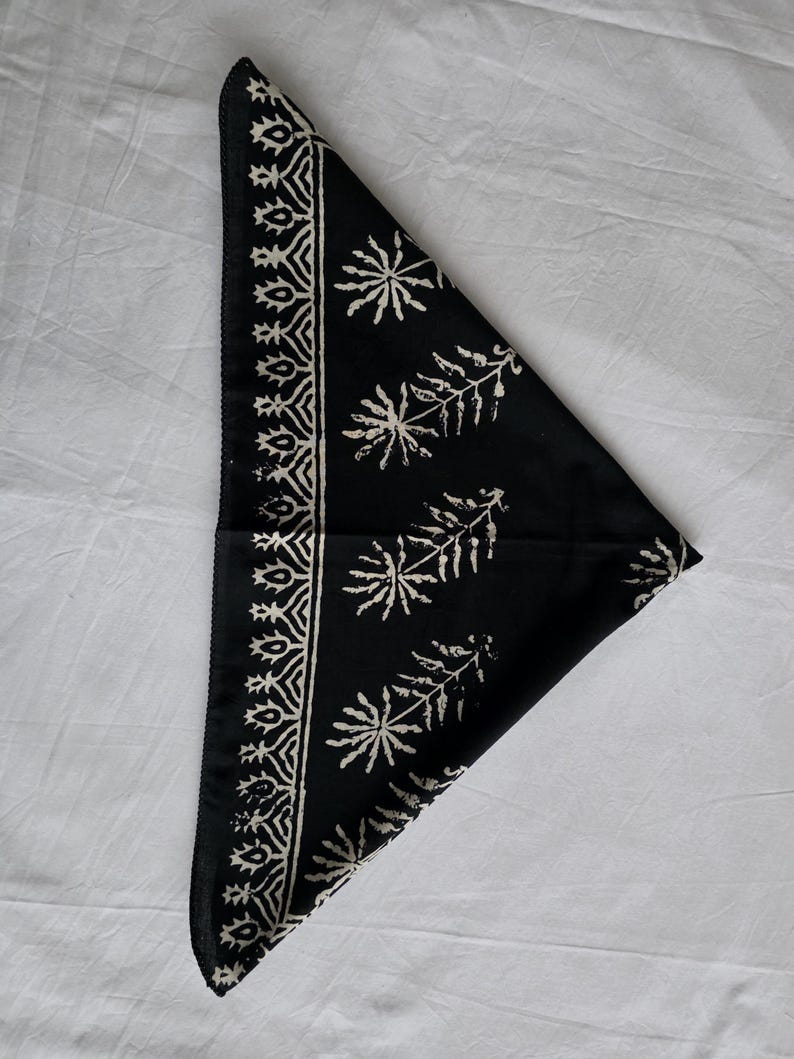 May include: Black bandana with a white floral and geometric pattern. The bandana is folded in a triangle shape, with the patterned border along one edge and floral designs across the surface. The fabric appears to be cotton or a similar material.