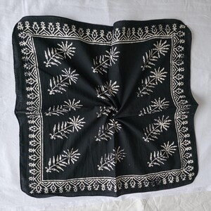 May include: Black square bandana with a white leaf and geometric pattern. The bandana has a decorative border and a central design of stylized leaves. The fabric is folded, creating a textured appearance.