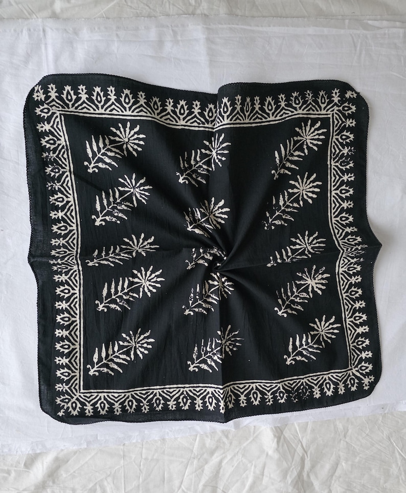 May include: Black square bandana with a white leaf and geometric pattern. The bandana has a decorative border and a central design of stylized leaves. The fabric is folded, creating a textured appearance.