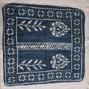 May include: A square, indigo-dyed textile with a white floral and geometric pattern. The design includes stylized leaves, floral motifs, and a border of star-like shapes. The fabric is likely cotton or a similar natural fiber.