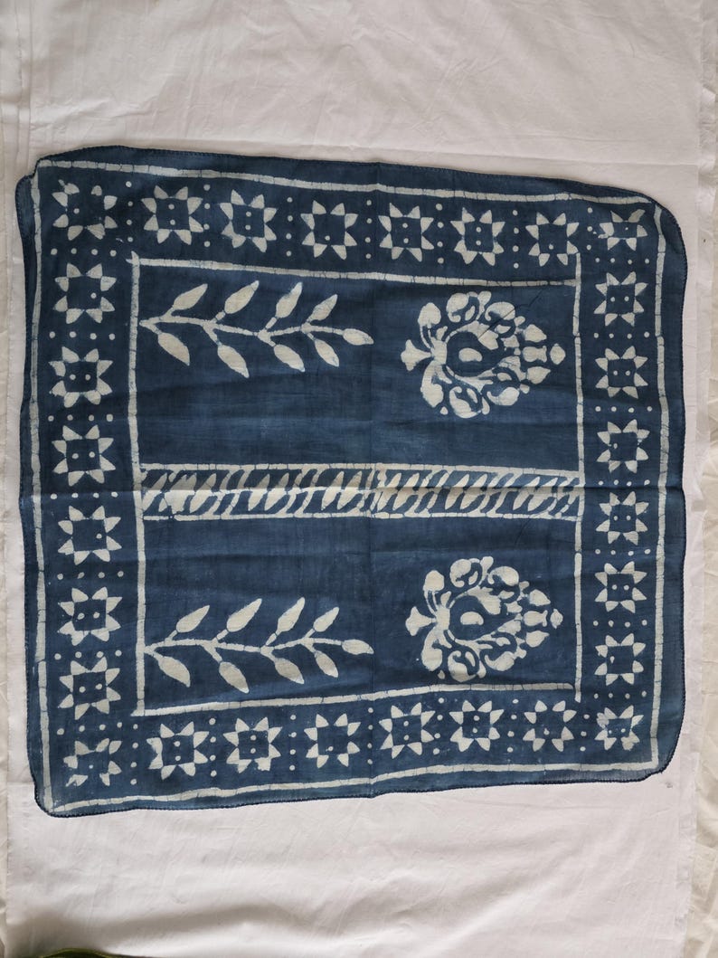 May include: A square, indigo-dyed textile with a white floral and geometric pattern. The design includes stylized leaves, floral motifs, and a border of star-like shapes. The fabric is likely cotton or a similar natural fiber.