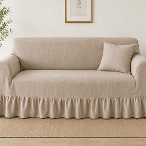 May include: A beige sofa cover featuring a textured fabric finish and a gathered ruffle skirt along the bottom edge. The cover is designed to fit over the back, seat, and arms of a sofa, and it includes a matching square throw pillow.