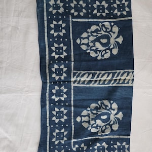 May include: A folded indigo blue textile with white floral and geometric patterns. The design includes a border of star-like shapes and central floral motifs. The fabric appears soft and likely made of natural fibers.