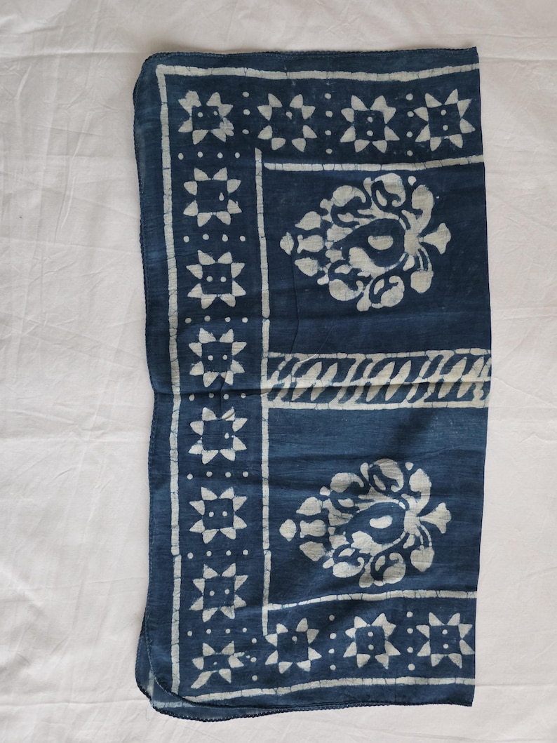 May include: A folded indigo blue textile with white floral and geometric patterns. The design includes a border of star-like shapes and central floral motifs. The fabric appears soft and likely made of natural fibers.