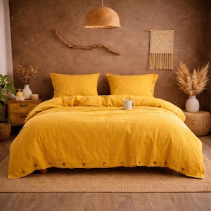 Può includere: Un set di biancheria da letto giallo senape con due federe abbinate, un copripiumino e un lenzuolo con angoli. Il copripiumino ha bottoni in legno sul bordo inferiore. Una tazza bianca è appoggiata sul letto. La camera da letto ha una combinazione di colori neutri.