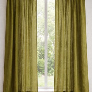 May include: Two olive green linen curtains hang on a white window frame. The curtains are floor-length and slightly gathered at the top. The window reveals a view of green trees and foliage, creating a natural backdrop.