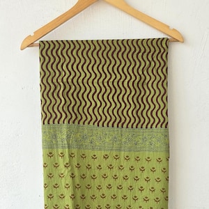 May include: A green and brown patterned fabric hanging on a wooden hanger. The top portion features vertical wavy lines in brown against a green background. The bottom has a floral and dot design.