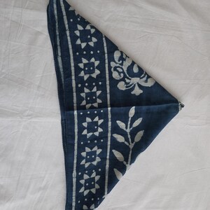 May include: A navy blue bandana with white geometric and floral patterns. The bandana is folded into a triangle, showcasing the detailed designs. The fabric appears to be soft and the patterns are evenly spaced.