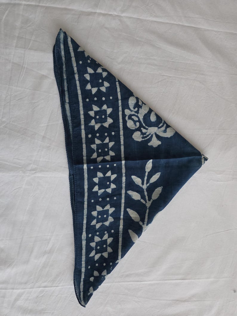 May include: A navy blue bandana with white geometric and floral patterns. The bandana is folded into a triangle, showcasing the detailed designs. The fabric appears to be soft and the patterns are evenly spaced.