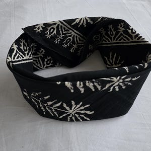 May include: Black headband with a white floral pattern. The fabric is folded over, creating a thick band. The headband is likely for fashion or to keep hair out of the face.
