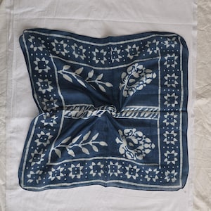 May include: A square, indigo blue scarf with a white floral and geometric pattern. The scarf features a central floral design and a border of star-like shapes. The fabric is folded, creating a textured appearance.