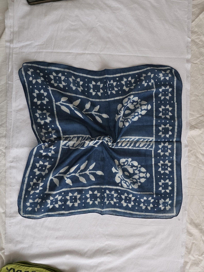 May include: A square, indigo blue scarf with a white floral and geometric pattern. The scarf features a central floral design and a border of star-like shapes. The fabric is folded, creating a textured appearance.