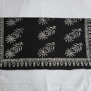 May include: A rectangular black textile with a white floral pattern. The design includes a border with a repeating geometric pattern and stylized leaf-like shapes scattered across the center. The textile is likely a decorative item.