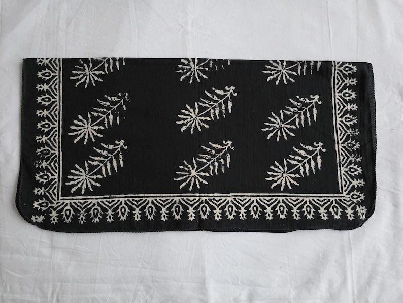 May include: A rectangular black textile with a white floral pattern. The design includes a border with a repeating geometric pattern and stylized leaf-like shapes scattered across the center. The textile is likely a decorative item.