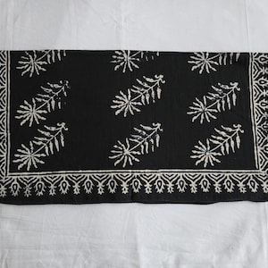 May include: A black rectangular textile with a white floral pattern. The design features stylized leaf and floral motifs, with a decorative border. The fabric appears to be soft and lightweight, suitable for a scarf or decorative cloth.
