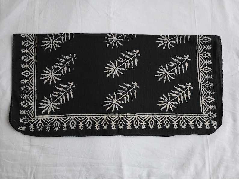 May include: A black rectangular textile with a white floral pattern. The design features stylized leaf and floral motifs, with a decorative border. The fabric appears to be soft and lightweight, suitable for a scarf or decorative cloth.