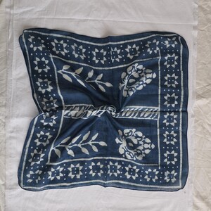 May include: A square, indigo blue textile with a white floral and geometric pattern. The fabric is folded, revealing the intricate design. The border features a repeating starburst motif, and the center has stylized floral elements.