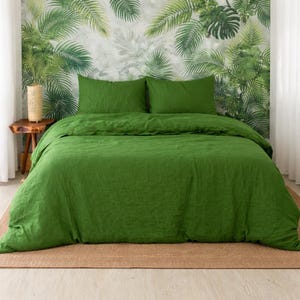 May include: A green linen bedding set, including a duvet cover and two pillowcases, is displayed on a bed. The bedding is a vibrant green color. The background features a tropical leaf pattern wallpaper. A small wooden side table with a lamp is visible.