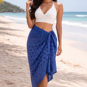 May include: A woman wearing a white halter top and a blue sarong. The sarong is wrapped around the waist and features a dark blue floral pattern. The background shows a beach with sand and ocean.