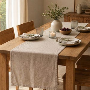 Op de afbeelding: Een lange, rechthoekige beige linnen tafelloper gedrapeerd over een houten eettafel. De loper heeft een getextureerd, geweven uiterlijk en ligt in het midden van de tafel, die is gedekt met borden, servetten, glaswerk en een vaas.