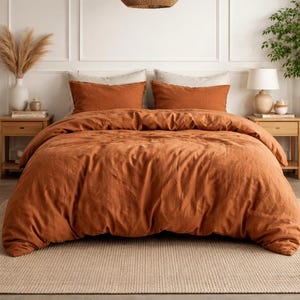 Puede incluir: Una cama con ropa de cama de color óxido, que incluye una funda nórdica y fundas de almohada. La cama está sobre una alfombra de color neutro. Dos mesitas de noche de madera flanquean la cama, cada una con una lámpara y objetos decorativos.