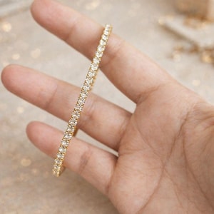 May include: A gold-colored tennis bracelet with numerous small, clear gemstones set closely together. The bracelet is held in a hand, showcasing its design and sparkle. The background is softly blurred, highlighting the jewelry.