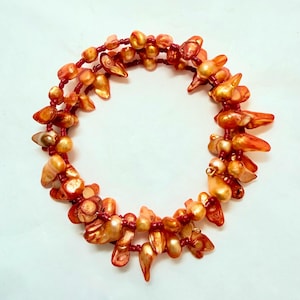 Op de afbeelding: Een ketting met oranje en gouden schelpvormige kralen en kleine rode kralen. De ketting is in een cirkelvorm gerangschikt, waarbij de schelpkralen in grootte en vorm variëren. De kralen hebben een glanzende afwerking, wat een aantrekkelijk ontwerp creëert.