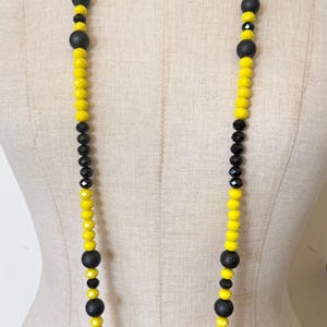 Può includere: Una lunga collana con sezioni alternate di perline gialle e nere. La collana ha un design simmetrico, con perline nere opache più grandi intervallate nelle sezioni di perline gialle. La collana è esposta su un manichino.