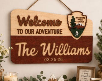 May include: A rectangular wooden wall sign with a rope hanger, featuring the text "Welcome TO OUR ADVENTURE" and "The Williams" with the date "03.25.26". It includes a National Park Service emblem with a mountain and Bigfoot graphic.