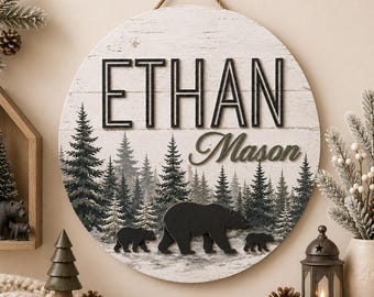 May include: A circular wooden wall sign featuring a white distressed wood grain background with the black text ETHAN and the script text Mason. The design includes a printed forest of pine trees and three dimensional black bear silhouettes.