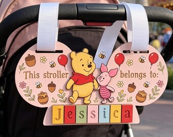 May include: A light pink stroller sign featuring Winnie the Pooh and Piglet, with the text 'This stroller belongs to:' and the name 'Jessica' on colorful blocks. The sign is decorated with bees, balloons, and acorns, and hangs from white straps.