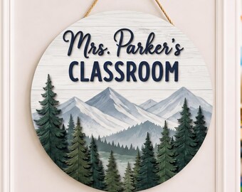 May include: A round wooden hanging sign featuring a white wood grain background, a mountain range illustration, and a forest of green pine trees. The text "Mrs. Parker's CLASSROOM" is printed in dark blue lettering. It hangs from a tan rope.
