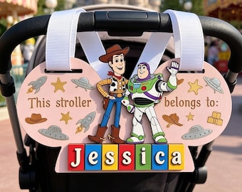 May include: A light pink stroller sign featuring Woody and Buzz Lightyear, with the text 'This stroller belongs to:' and the name 'Jessica' displayed on colorful blocks. The sign is decorated with stars, rockets, and flying saucers, attached by white straps.