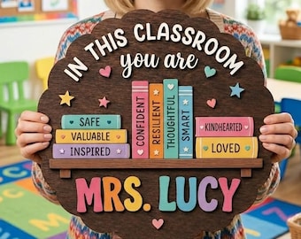 May include: A dark brown scalloped wooden sign featuring the text IN THIS CLASSROOM you are, with colorful book illustrations labeled SAFE, VALUABLE, INSPIRED, CONFIDENT, RESILIENT, THOUGHTFUL, SMART, KINDHEARTED, LOVED, and the name MRS. LUCY.