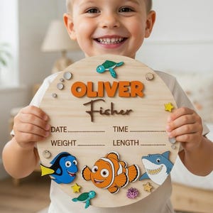 May include: A circular wooden baby birth announcement sign featuring the name Oliver Fisher in orange and brown text. The board includes blank lines for date, time, weight, and length, decorated with colorful wooden cutouts of sea creatures.