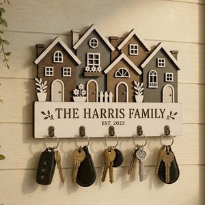 Può includere: Un portachiavi da parete in legno con quattro facciate di case in miniatura nei toni del marrone e del grigio. La base bianca riporta la scritta THE HARRIS FAMILY EST. 2023 e presenta cinque ganci in metallo con vari mazzi di chiavi.