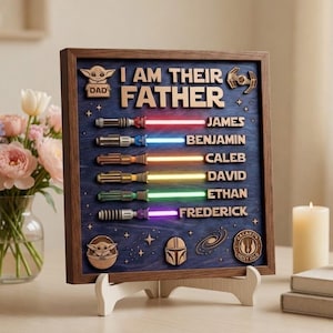Może przedstawiać: Drewniany szyld w ramie z napisem "I AM THEIR FATHER" i spersonalizowanymi imionami. Zawiera wzory mieczy świetlnych z kolorowymi wiązkami. Zawiera ikony z motywem Gwiezdnych Wojen i drewniany stojak.