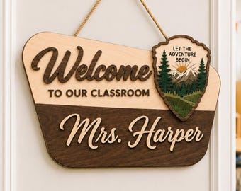 May include: A trapezoidal wooden classroom sign with a light wood top and dark wood bottom. It features the text "Welcome TO OUR CLASSROOM" and "Mrs. Harper" in dark script. A shield-shaped emblem shows a mountain, trees, and "LET THE ADVENTURE BEGIN".