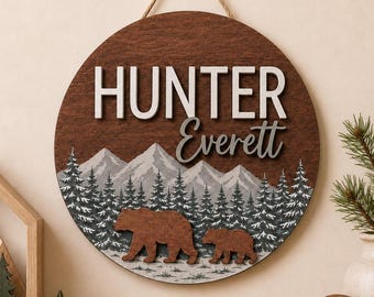 May include: A circular wooden wall sign featuring the name Hunter Everett in white and grey lettering. The design includes a layered mountain range, a forest of pine trees, and two brown bear silhouettes hanging from a jute rope.