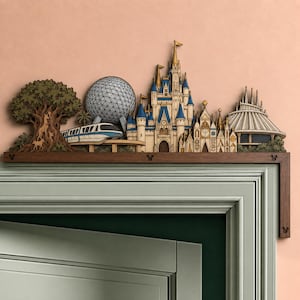 Op de afbeelding: Een houten deurdecoratie met een gelaagd, lasergesneden ontwerp van een pretpark-skyline. Het bevat een grote boom, een geodetische bol, een monorail, een kasteel met blauwe torenspitsen en een kegelvormige structuur op een donkerhouten lijst.
