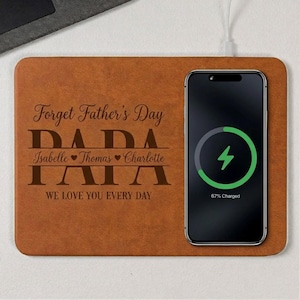 May include: A brown leather wireless charging pad with the text "Forget Father's Day" and "DADA" engraved. A black smartphone is charging on the pad, displaying a charging symbol and "67% Charged". The names Isabelle, Thomas, and Charlotte are also engraved.